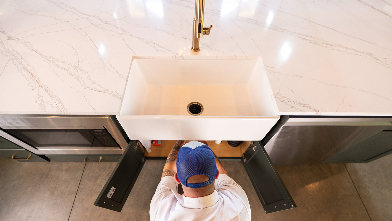 A plumber in a blue uniform inspects a garbage disposal and plumbing connections under a kitchen sink.