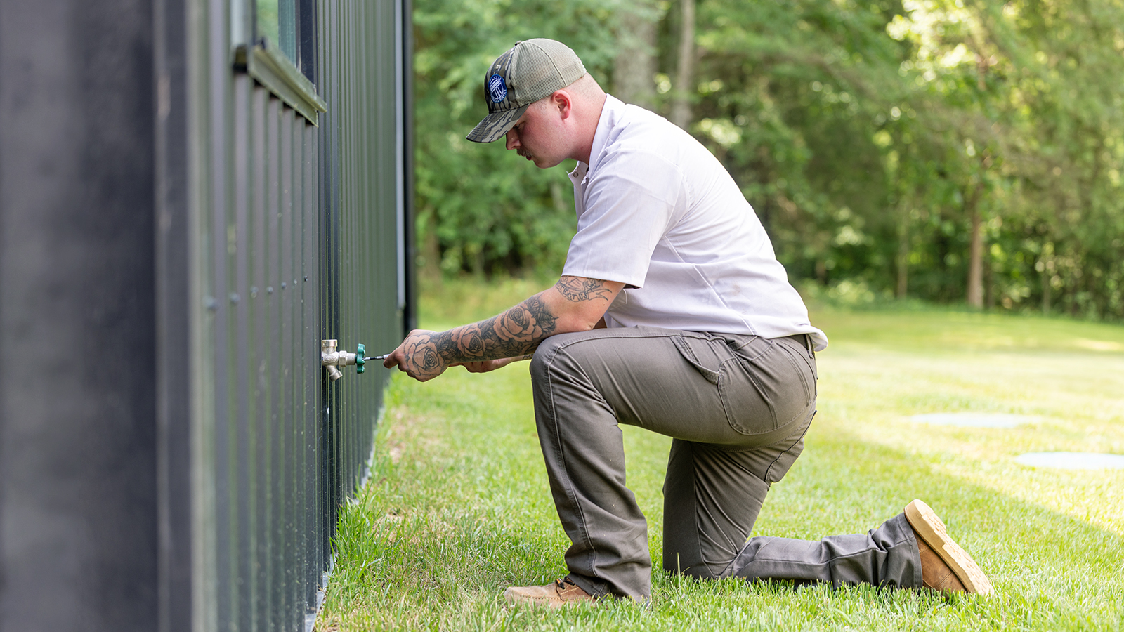A plumber in a blue uniform uses pliers to service an outdoor spigot on the exterior of a home.