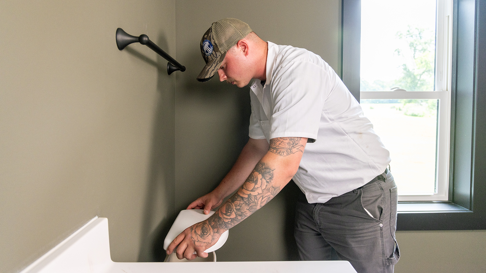 Plumber wearing a blue sweatshirt and cap working on the inside of a toilet tank in a home bathroom.