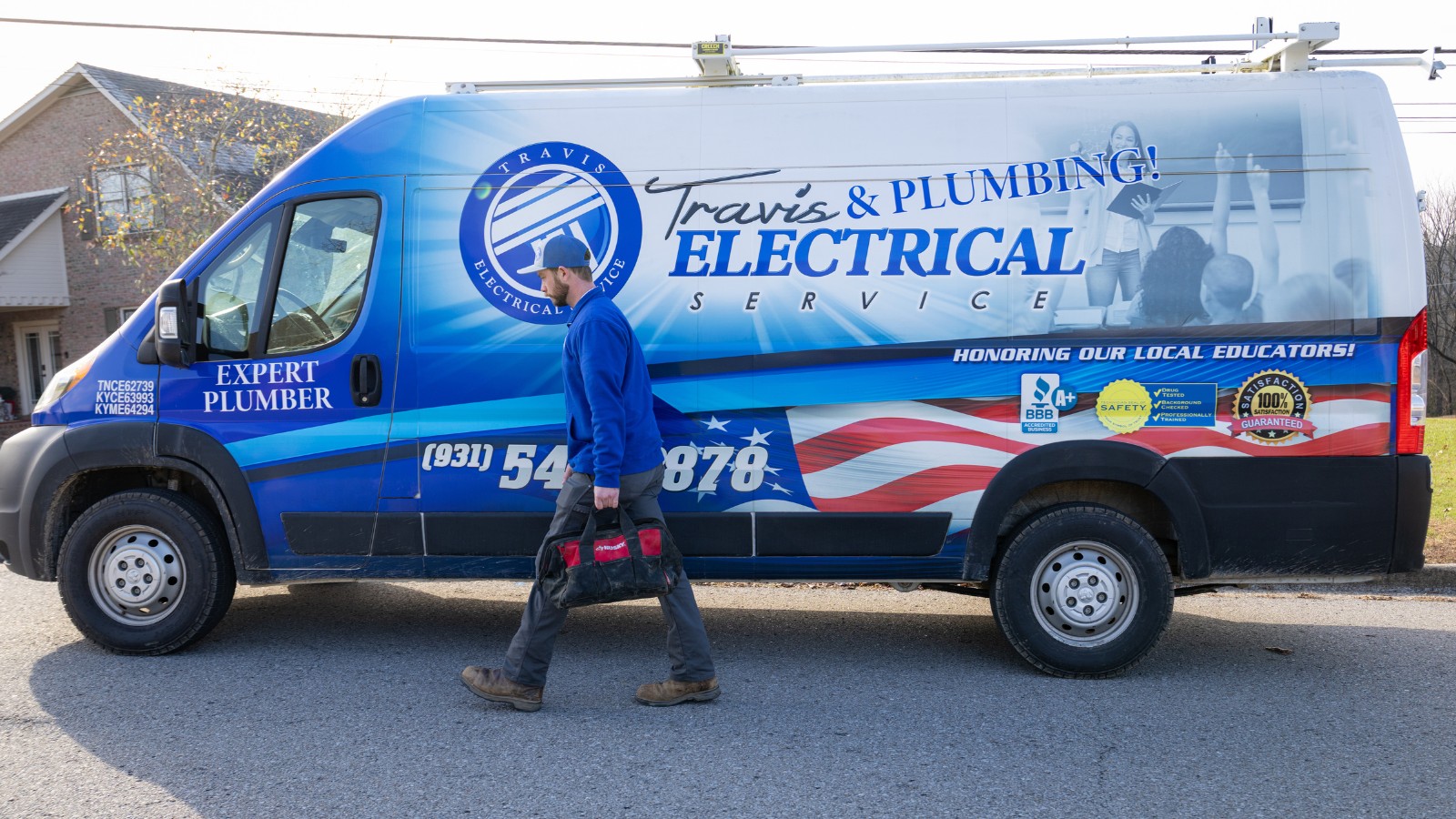 Plumber with blue uniform walking with tool bag in hand in front of a Travis Electrical Service & Plumbing truck.